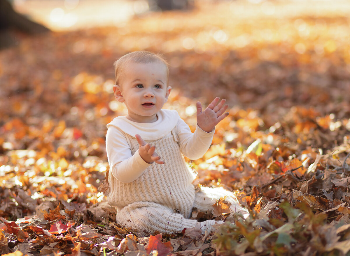 Baby in beige romper is sitting in the leaves for a fall family photoshoot. Captured by Brooklyn family photographer Chaya Bornstein