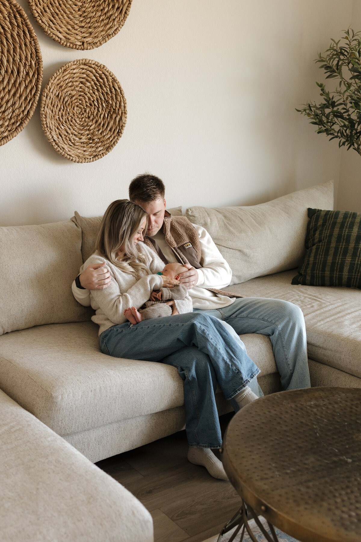 Husband and wife sitting on tan couch holding newborn looking down and touching the babys face during in home newborn session in longmont colorado