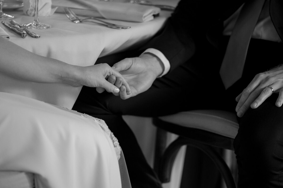 A tender black and white wedding photo of a couple holding hands during emotional toasts, capturing love and connection.
