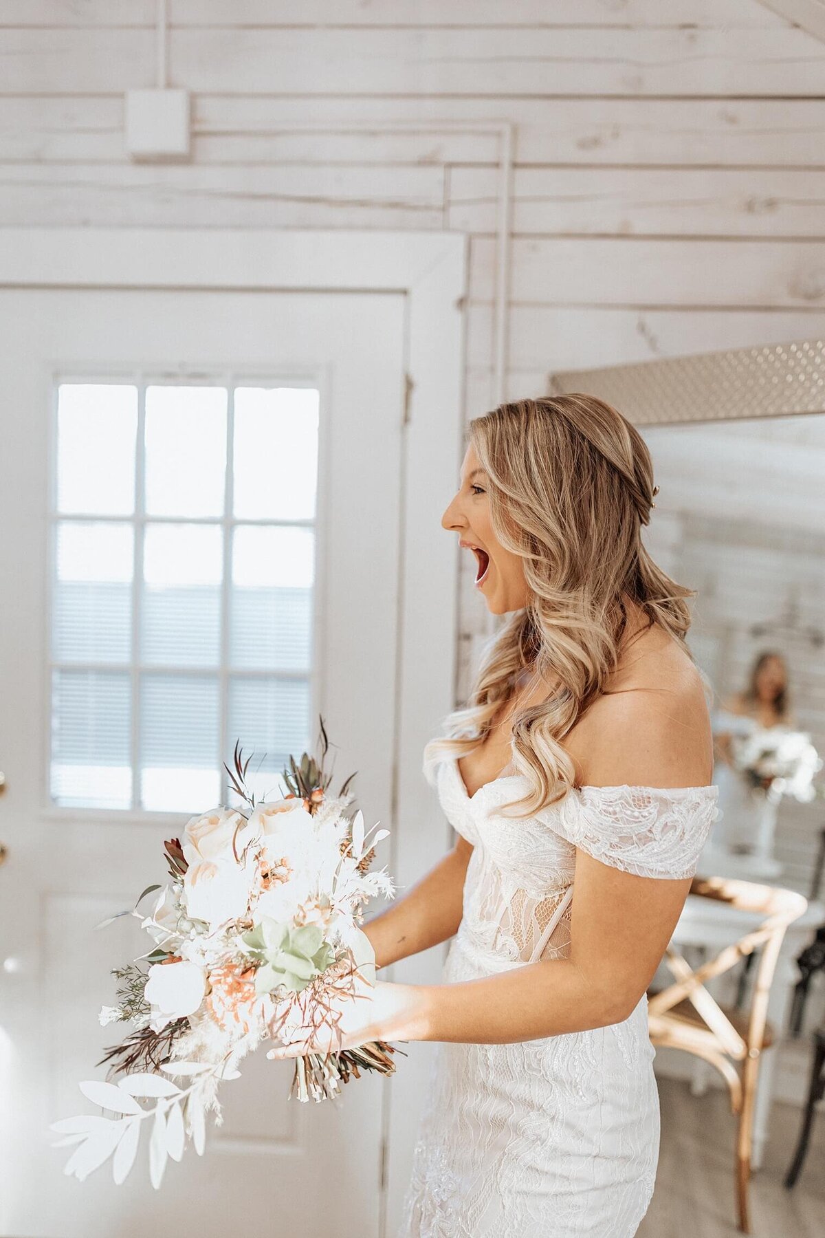Women in white wedding dress holding bouquet smiling as she looks at herself in the mirror