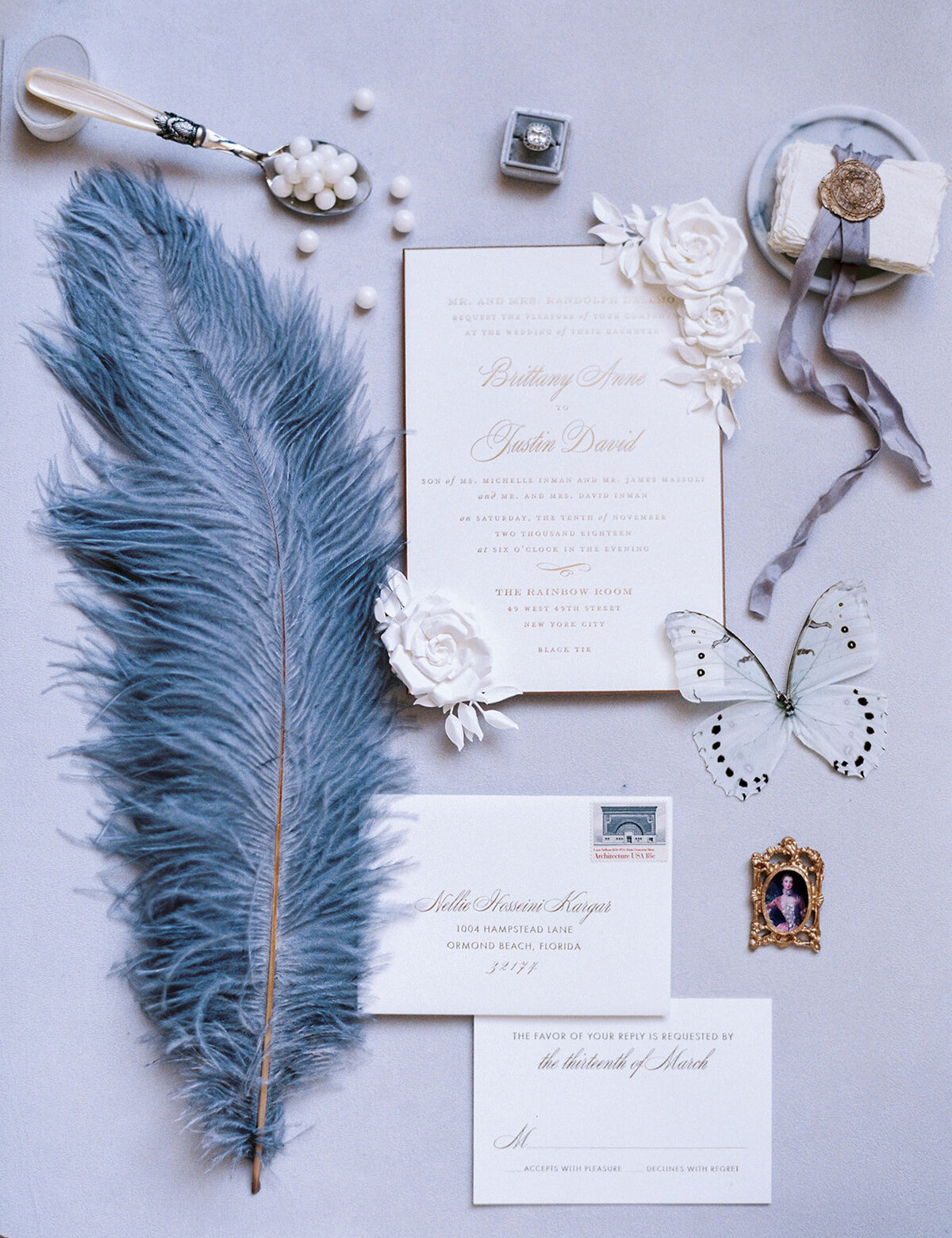 A flat lay photo of elegant wedding stationery featuring invitations, an RSVP card, and envelopes adorned with cursive text and floral designs. Surrounding the stationery are a blue feather, a decorative butterfly, a miniature portrait, and a bowl of white candies.