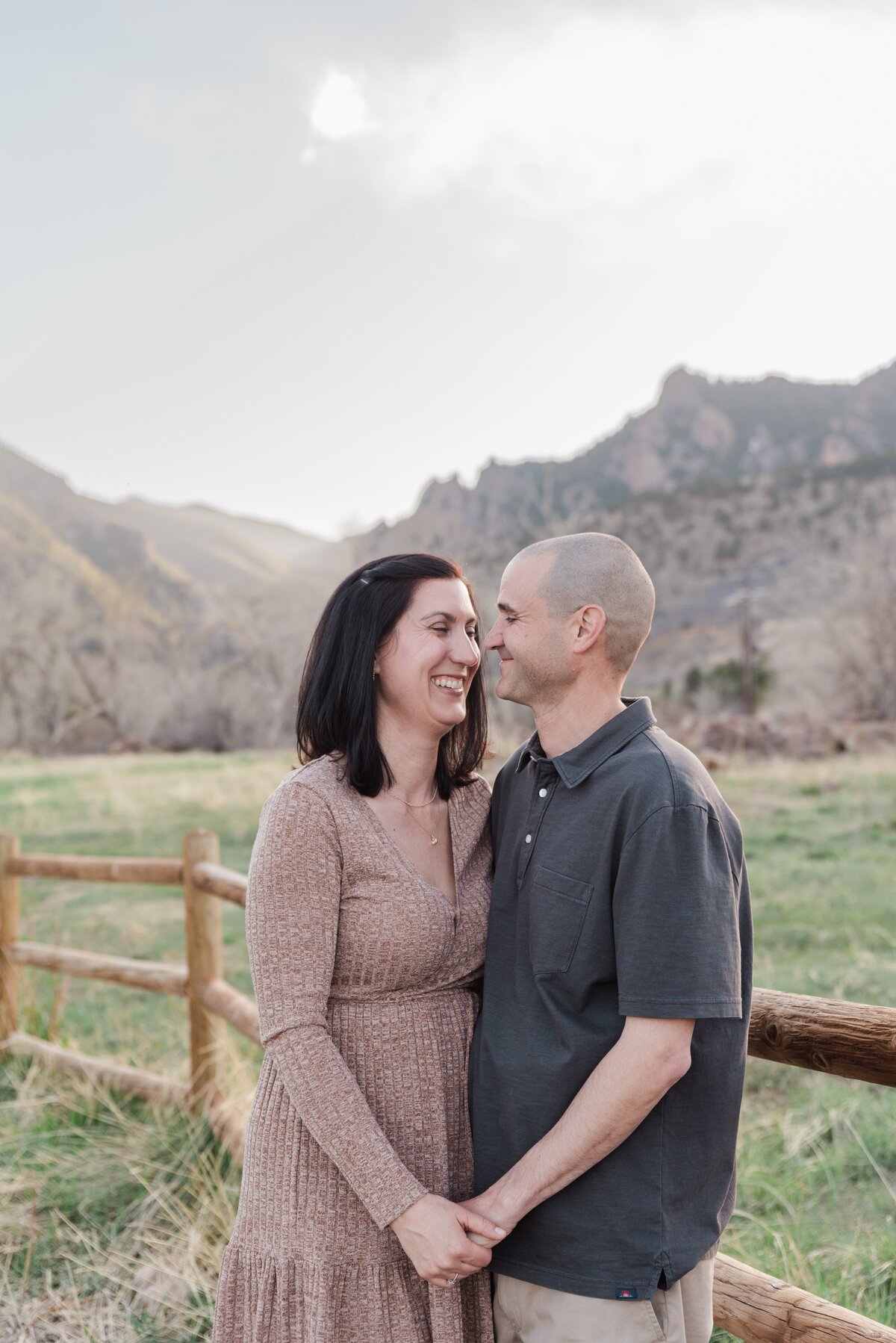 A woman in a brown long sleeve dress and a man in a dark grey polo hold hands and look at each other facing each other and smile