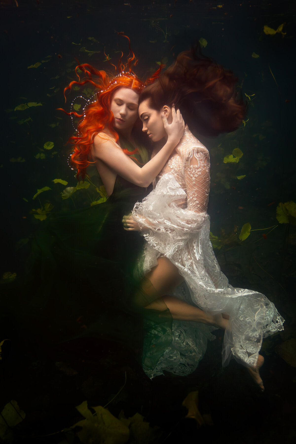 Two women float gracefully underwater, their arms intertwined in a delicate embrace. One woman with vibrant red hair wears a deep green gown, while the other is adorned in intricate white lace. Their peaceful expressions and flowing gowns create a mesmerizing underwater maternity photo, surrounded by drifting leaves and soft light. Dallas underwater photography captured with elegance and artistry.