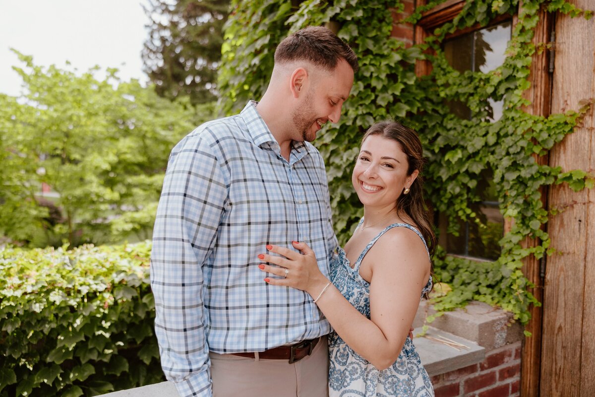 Engaged couple smiling during their Marist College engagement session, standing by ivy-covered brick and wood architecture in the Hudson Valley.