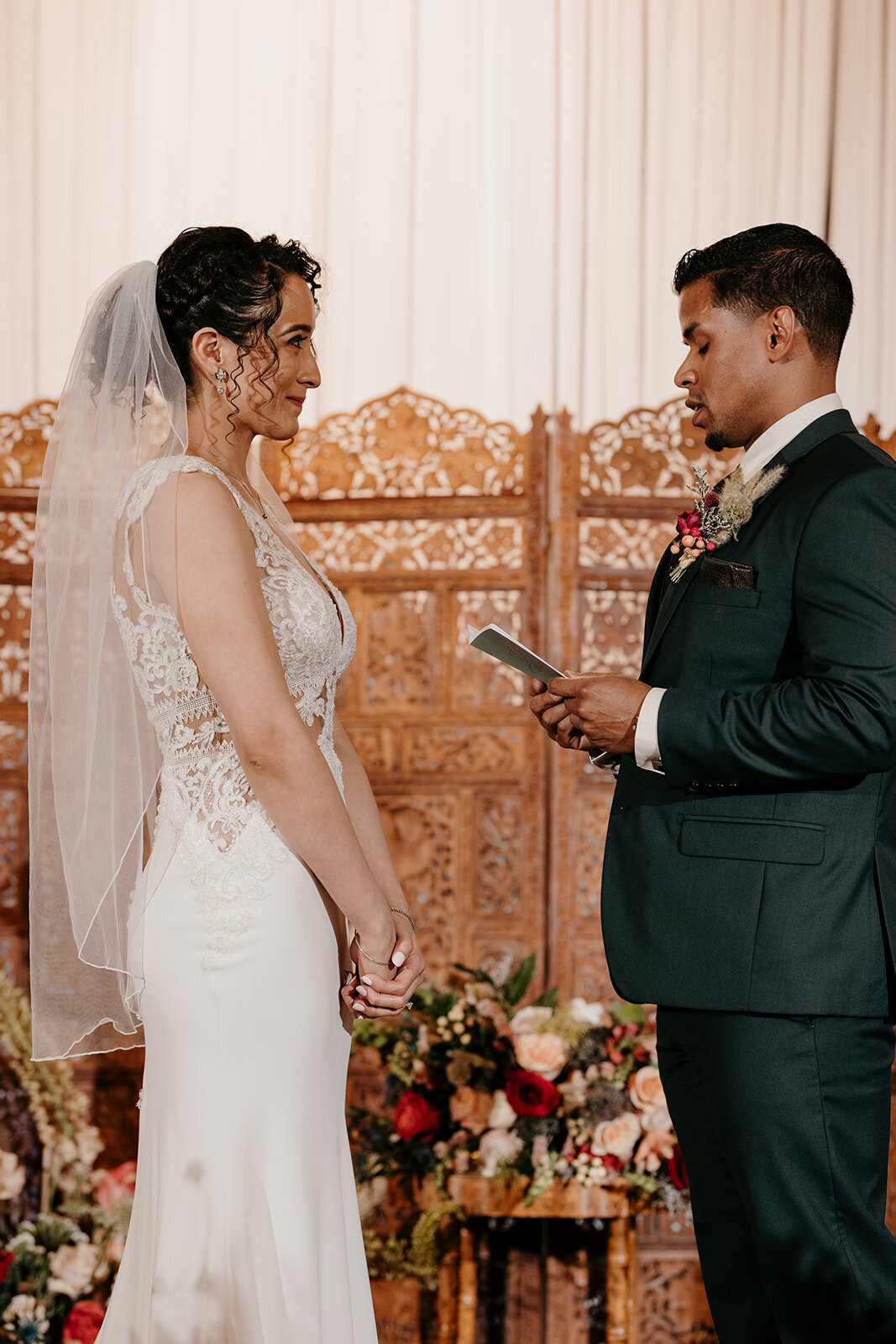 Bride and groom during their wedding ceremony, with the groom reading vows from a note while the bride smiles warmly, set against a decorative wooden backdrop and floral arrangements.