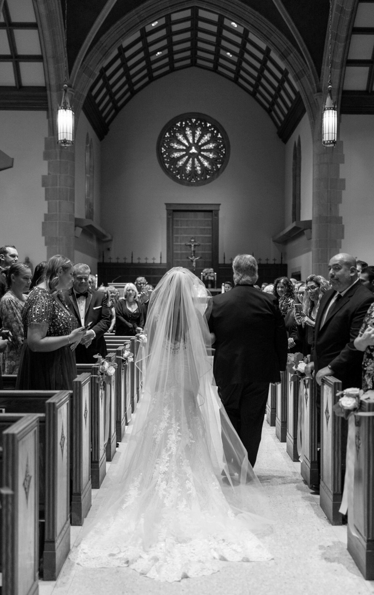 A black and white image of a wedding ceremony in a church. A bride in a long veil walks down the aisle with an older man, possibly her father. Guests on both sides watch the procession, and a large, circular stained-glass window is visible at the front.