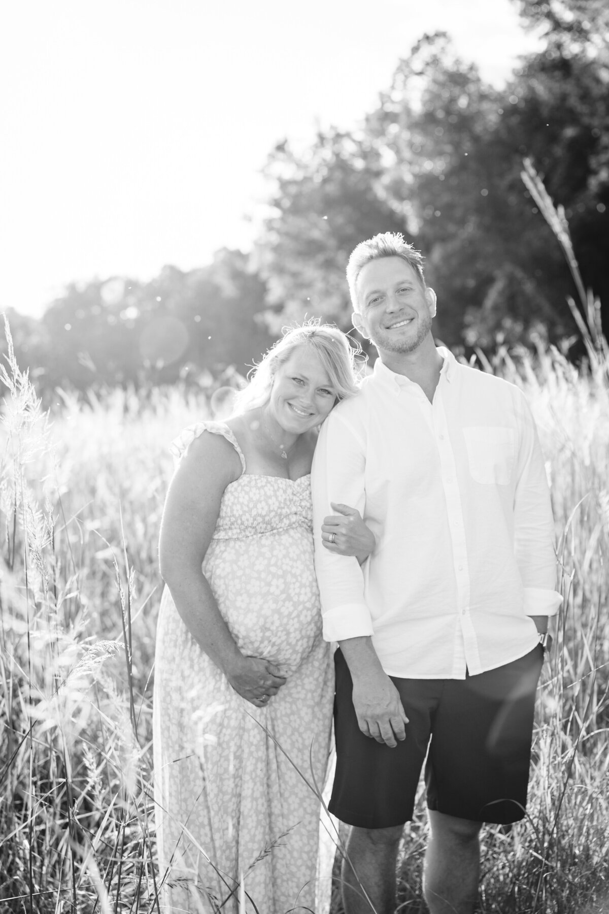 A black-and-white photo of a pregnant couple standing close together in a grassy field, the woman leaning her head on the man's shoulder.
