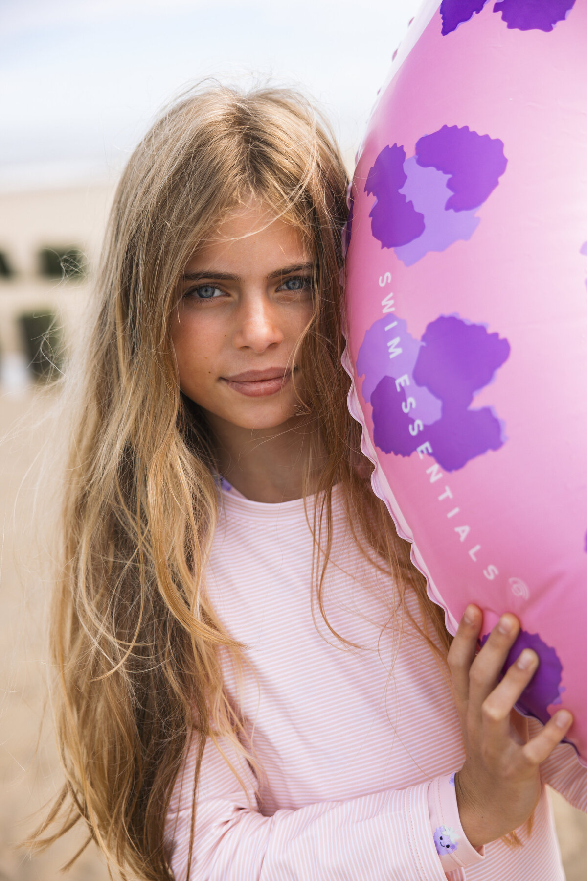 A girl with long blonde hair holds a pink leopard-print Swim Essentials inflatable, wearing a light pink striped rash guard on the beach.