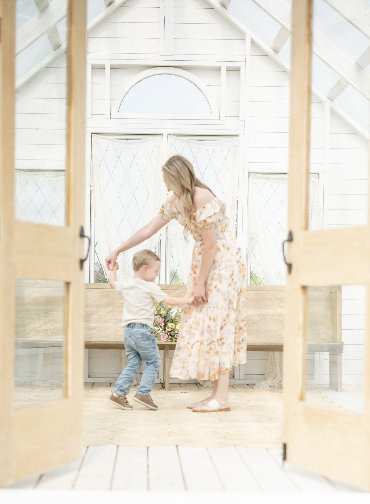 Mom and toddler playing together inside a bright conservatory with white wood and soft window light.