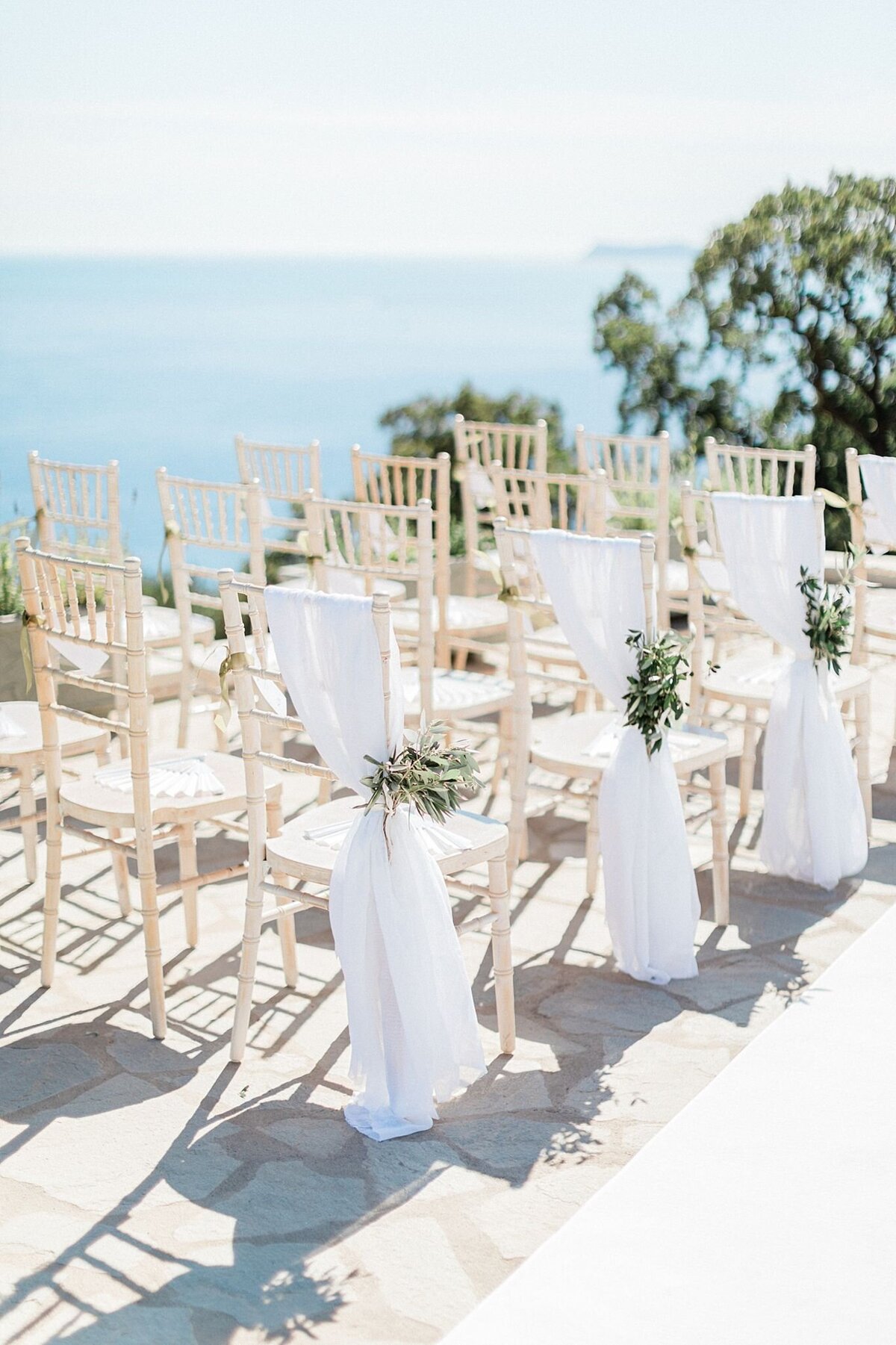 Outdoor seaside wedding ceremony setup with ivory chairs, white draping, and olive branch accents overlooking the Ionian coast