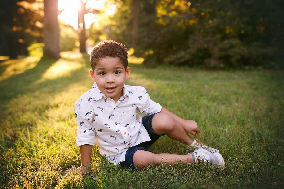 A-young-boy-smiling-while-sitting-in-the-grass-during-a-golden-hour-photo-session-in-the-Meridian-Kessler-neighborhood-on-the-north-side-of-Indianapolis