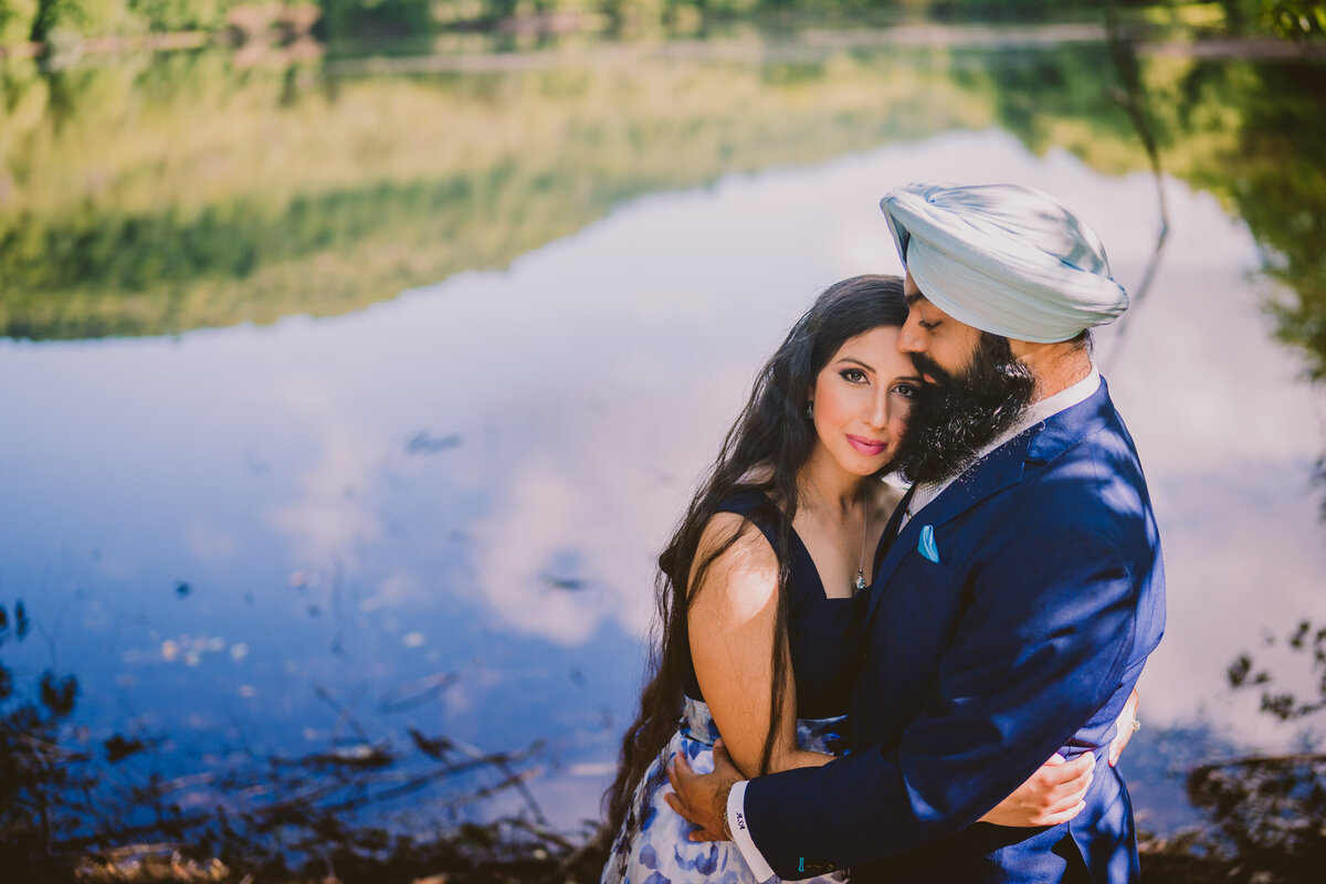 An engaged couple embrace in front of a lake at the ramapo valley reservation in nj