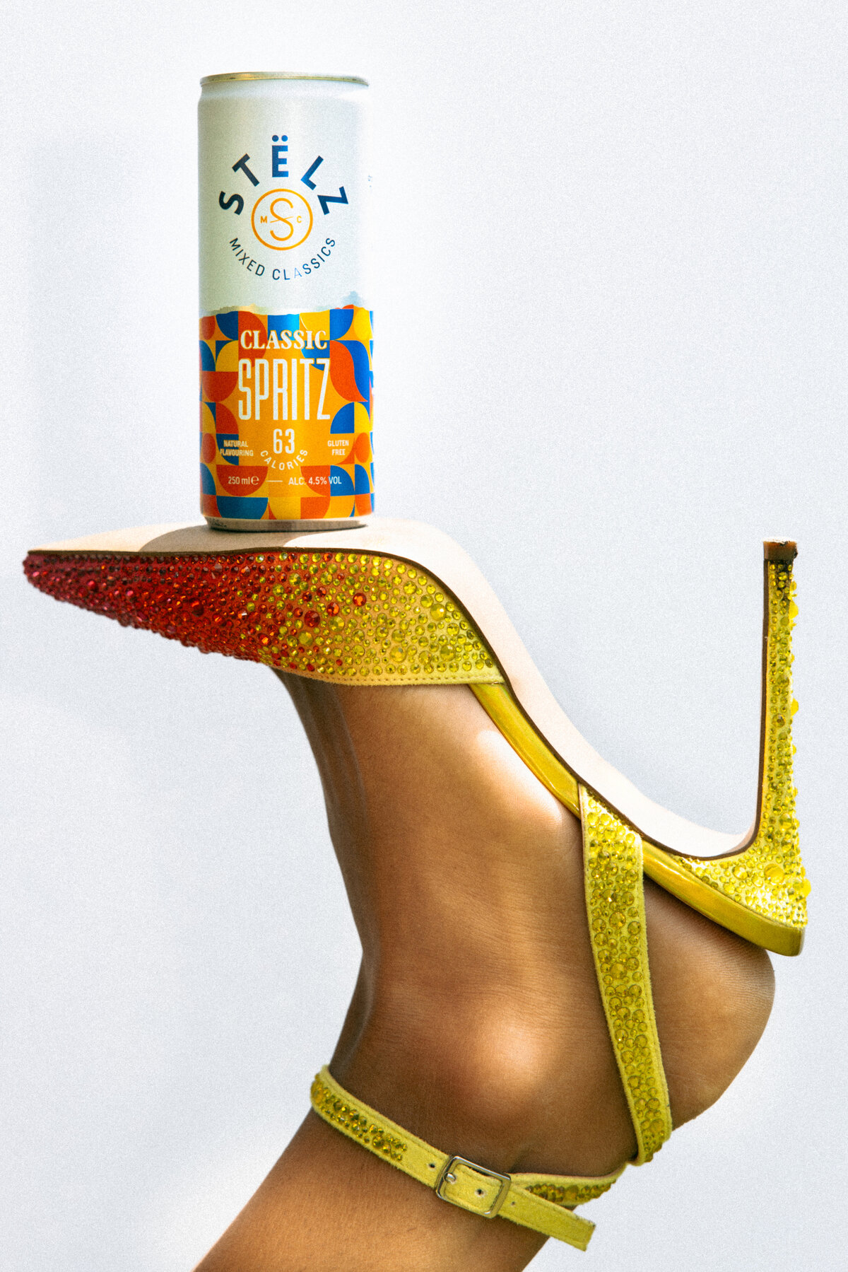 A close-up shot of a STËLZ Classic Spritz can elegantly balanced on the heel of a yellow, rhinestone-encrusted high heel. The image captures the fusion of style and refreshment, highlighting the product in a unique and creative way. Photography by Bo Eppink.