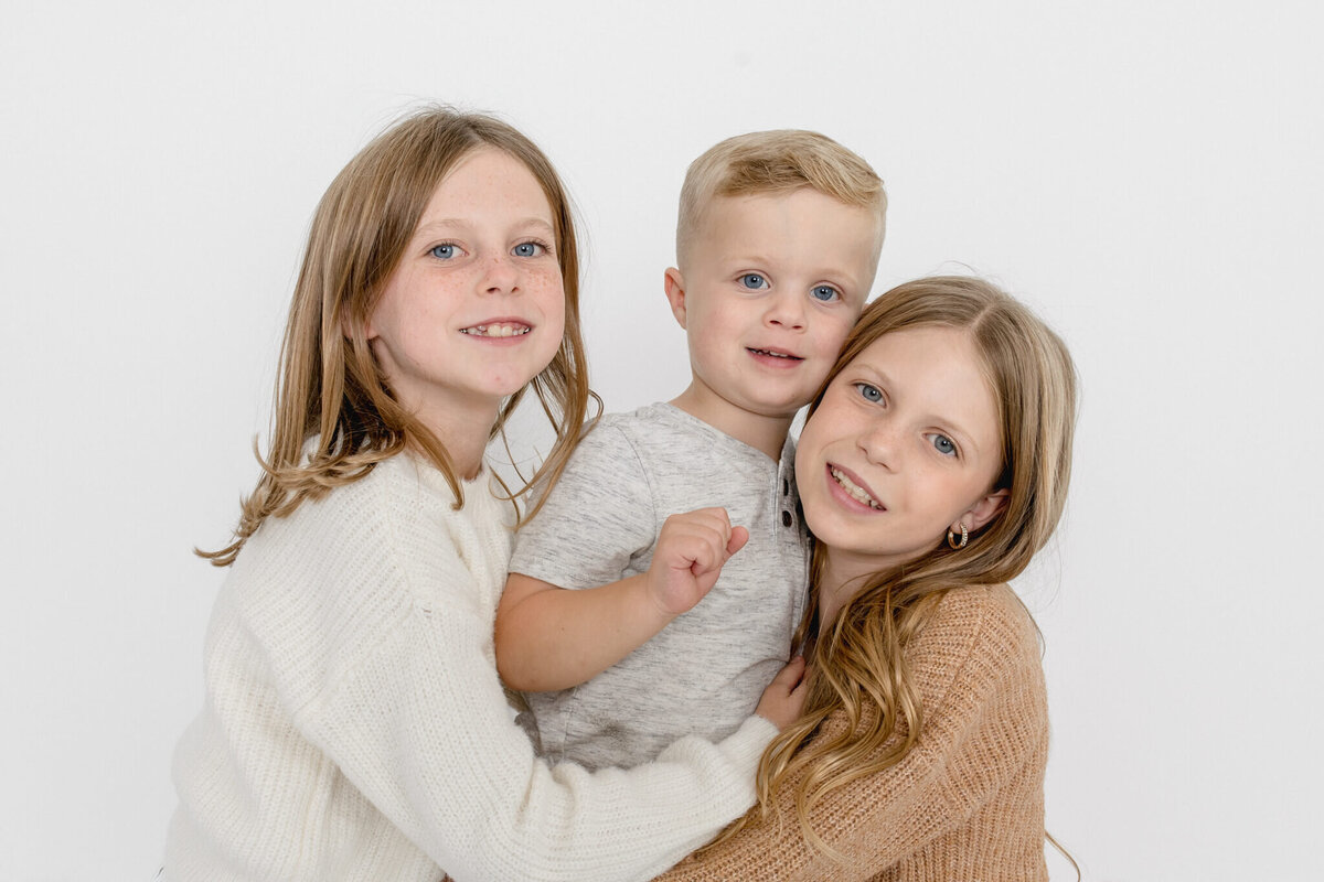 Two little girls hugging their little brother and smiling at the camera in a white studio in Tustin.