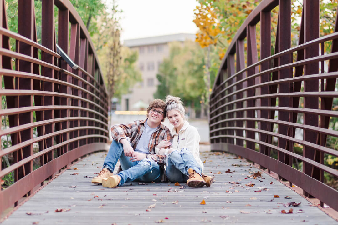 An engaged couple sit on a bridge smiling at the camera.