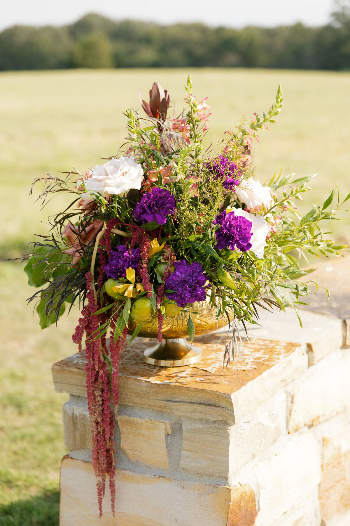 Vibrant mixed-floral wedding arrangement featuring white roses, purple blossoms, cascading amaranthus, and lush greenery styled in a gold compote bowl on a stone column outdoors.