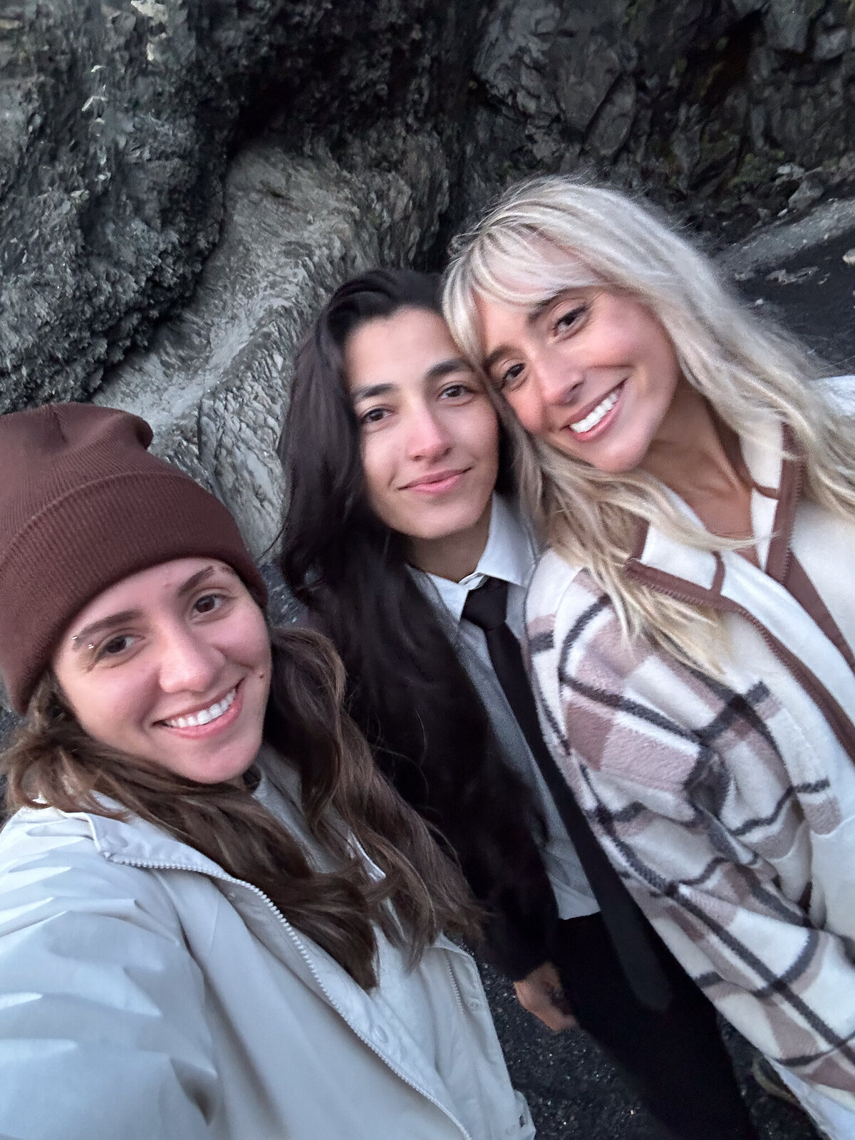 Female photographer smiling with content creator Sarah Carollyn and her girlfriend at Reynisfjara Beach, Iceland, wearing winter layers by the rocky coastline during a Destination Elopement.