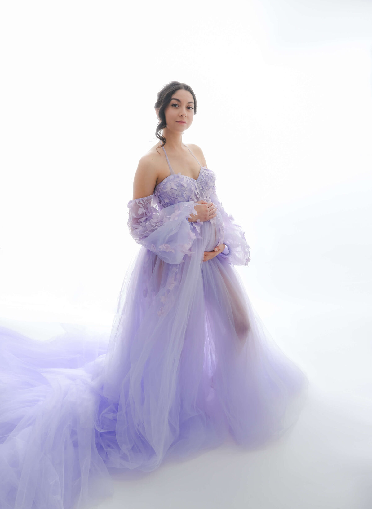 Expectant mother is wearing a floor length, lavender, off the should dress. She cradles her baby bump  in this pose during her maternity photo shoot. Image by Beth Benecki of E. Benecki Photography