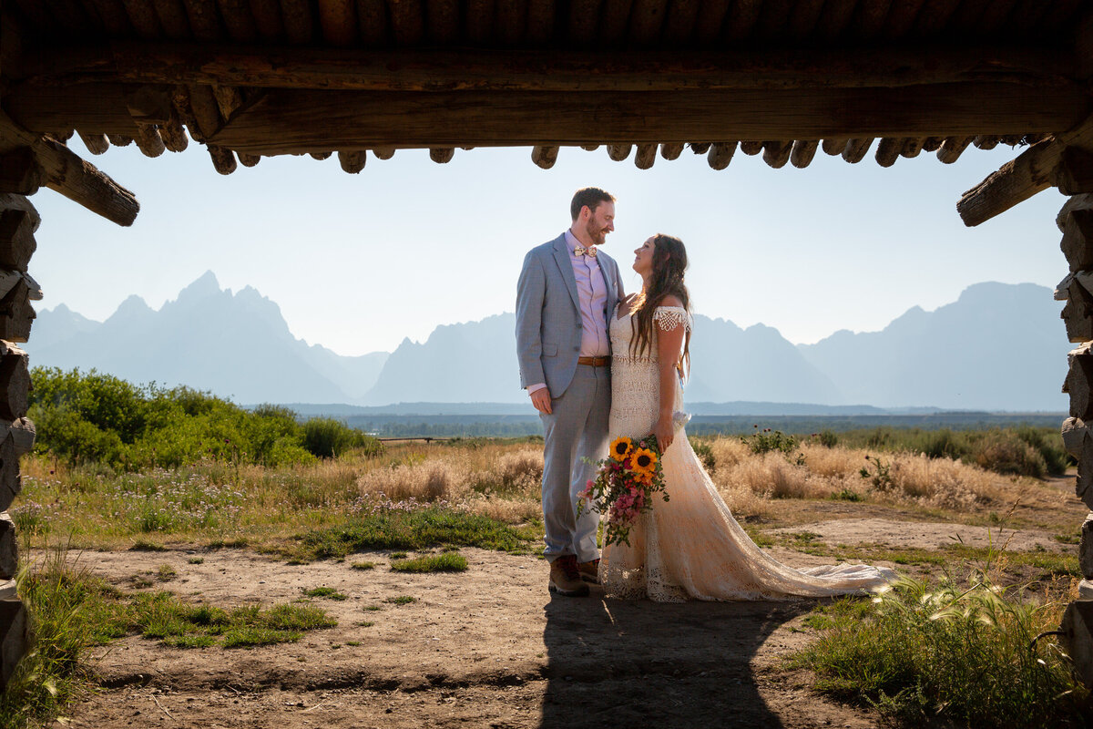 A bride and groom stand together on their wedding day as their Wyoming elopement photographer takes their picture through a barn.