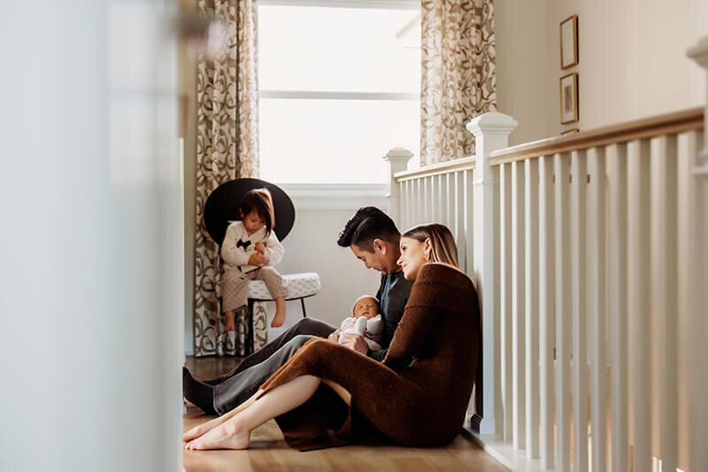 Mom and dad snuggle newborn baby sitting against stair rails while toddler daughter plays in the background during their at home newborn session in Denver