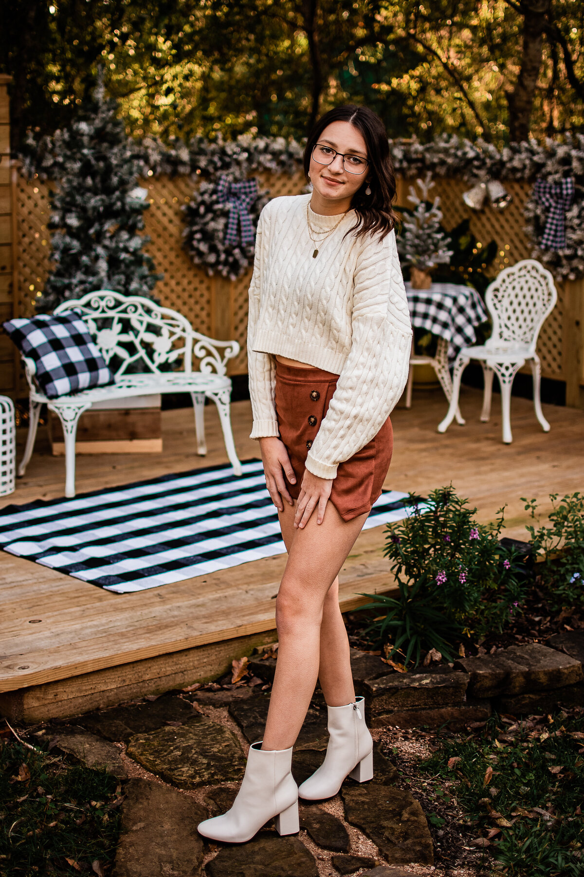 A senior wearing an orange skirt, white sweater, and white boots stands in front of a patio decorated with white and black plaid Christmas items.
