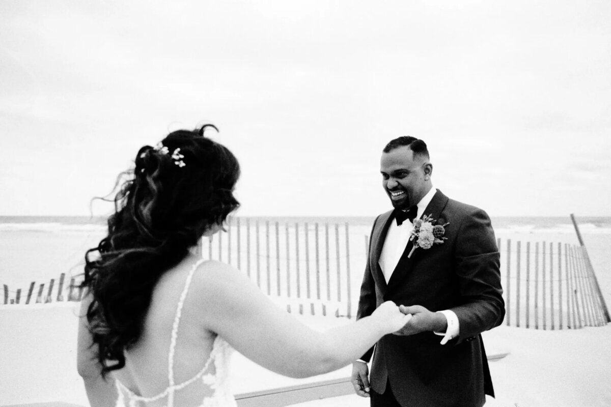 A bride and groom hold hands and smile at each other on a beach, waves crashing behind them and a wooden fence nearby—captured by a talented NJ wedding photographer. The bride faces the ocean, while the groom, in a suit and boutonniere, beams with joy.