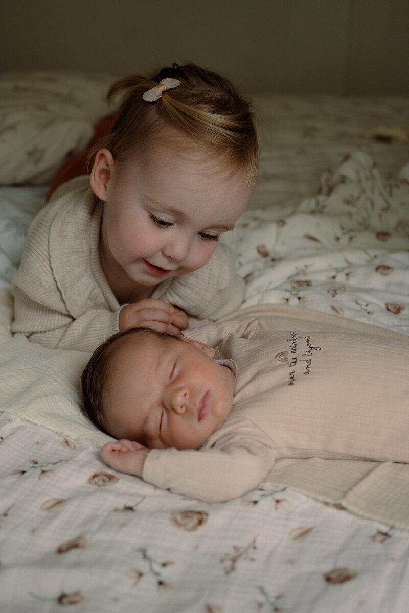 Grote zus kijkt liefdevol naar haar slapende kleine broertje tijdens een newbornshoot