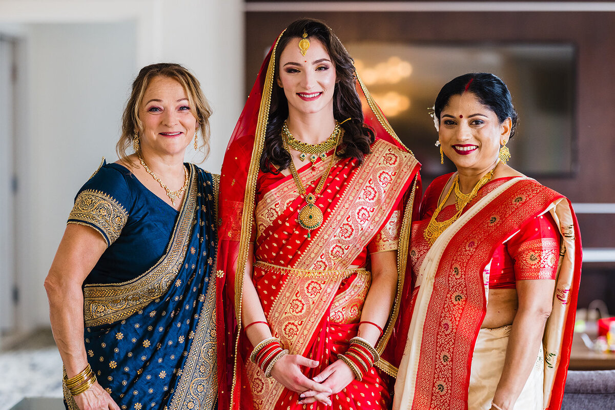 A bride in a red saree standing with two older women, one in blue and the other in red, all smiling and adorned in traditional Indian attire.