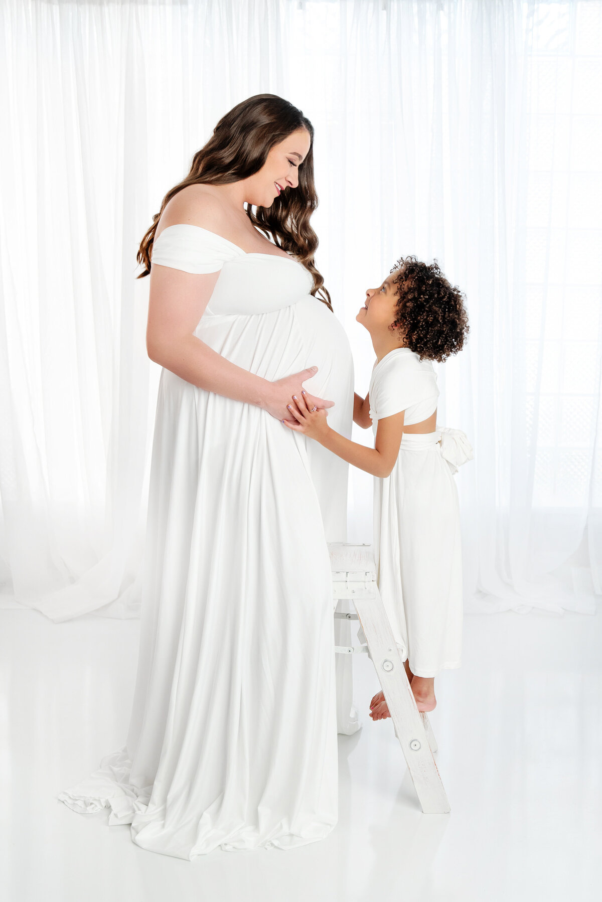st-louis-maternity-photographer-mother-and-child-smiling-at-eachother-holding-pregnant-belly-dressed-in-monocromatic-white-outfits-against-white-background