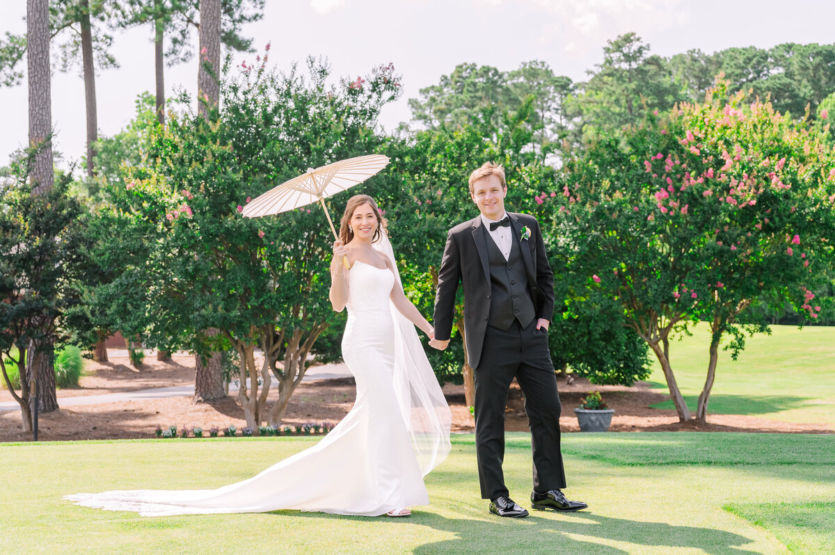 A happy bride and groom walking holding hands through a vividly green golf course in the summertime while holding a white umbrella as they enjoy their Raleigh wedding photos by JoLynn Photography