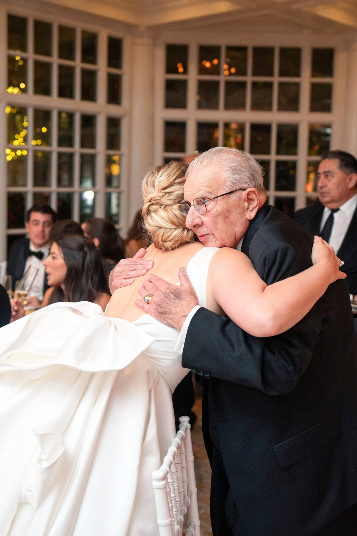 A bride in a white dress hugs an elderly man in a suit at a formal event. Guests are seated at tables in the background, surrounded by large windows.