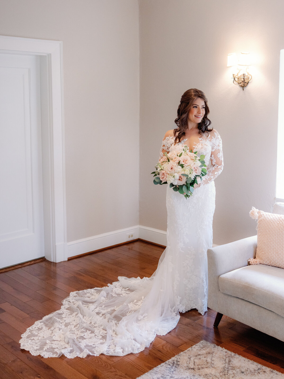 A bride, in her wedding dress and holding her blush pink and white bouquet, looks out the window of her getting ready space