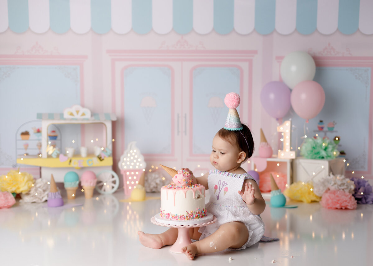 Ice cream shop themed cake smash in Palm Beach and Palm Beach Gardens photography studio. Baby girl is wearing an ice cream romper sitting behind a cake with a melted ice cream cone on top. In the background are soft pink and teal ice cream shop backdrop with pastel colored balloons and ice cream cones.
