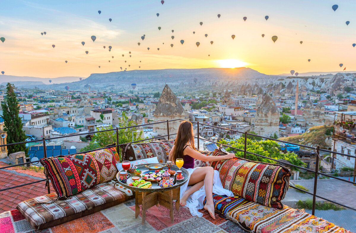 A woman sits on a colorful, cushioned terrace overlooking Cappadocia at sunrise. Hot air balloons fill the sky above a scenic landscape of rock formations and city rooftops. A round table holds a traditional Turkish breakfast spread.