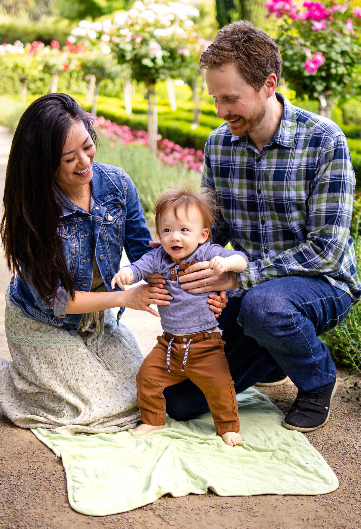Bay Area Family Portfolio - Ellobelle Photography shows proud parents helping baby stand on blanket in blooming garden.