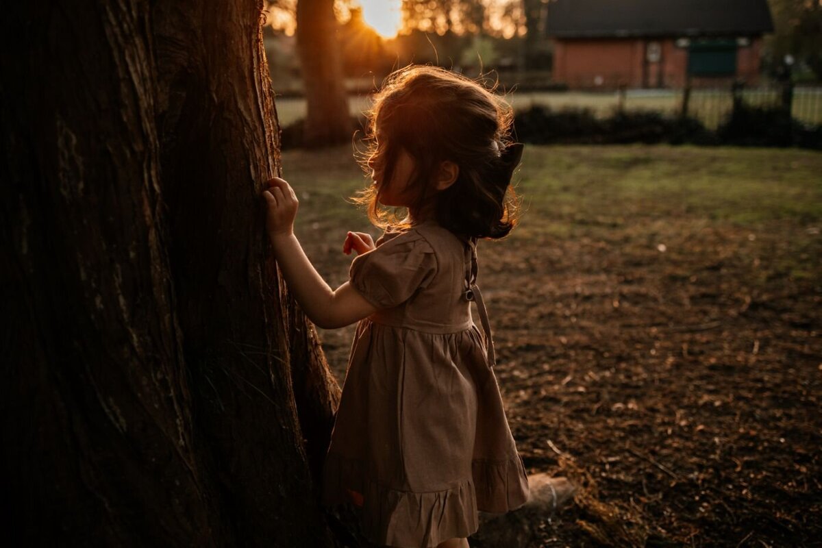 A young girl in a brown dress gently touches the bark of a large tree, as the setting sun casts a warm golden light in the park. Children's photography by Ruaa Hamdan