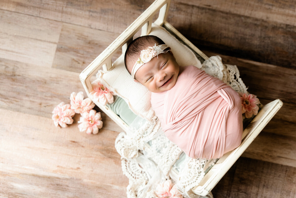 baby girl wrapped in pink smiles whiling sleeping in tiny cream bed with lace and flowers around