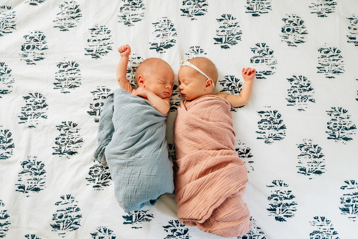 Two newborn twins lie side by side on a patterned bedspread, each wrapped in a soft blanket.