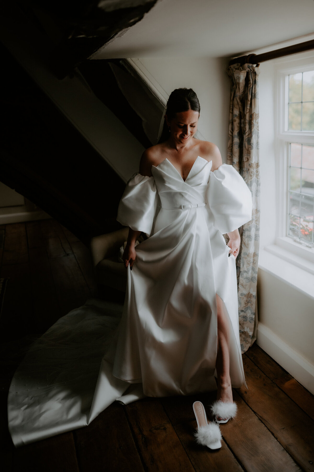 A bride slips into her shoes whilst standing in a window in her wedding dress and bell sleeves.