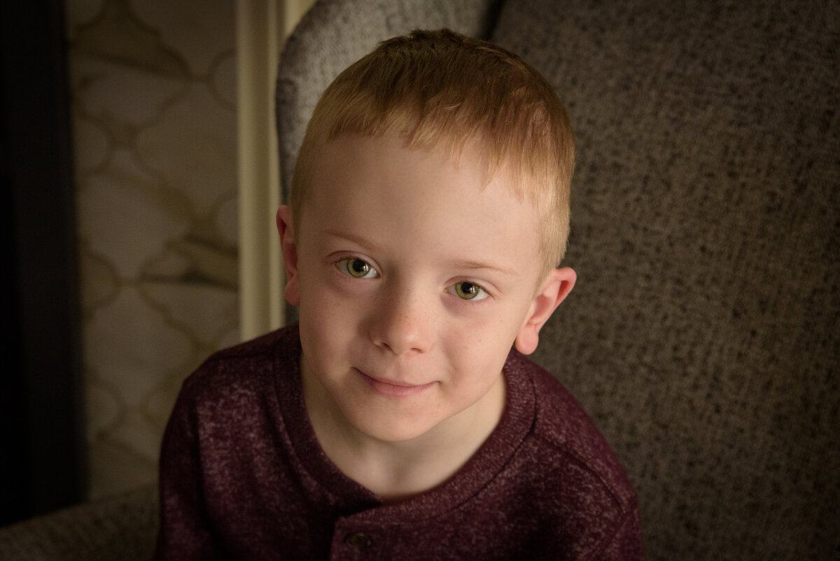 Natural light portrait of young boy wearing maroon shirt sitting in chair in his home in Green Bay, Wisconsin.