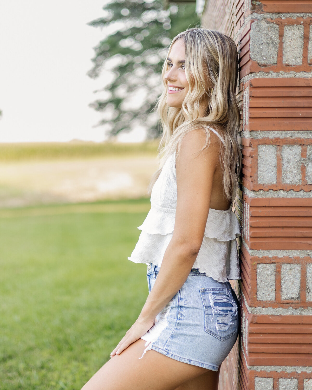 high school female senior with long blonde hair, standing in the grass, leaned against a brick wall, looking away from the camera smiling, wearing blue jean shorts, and a white spaghetti strapped shirt