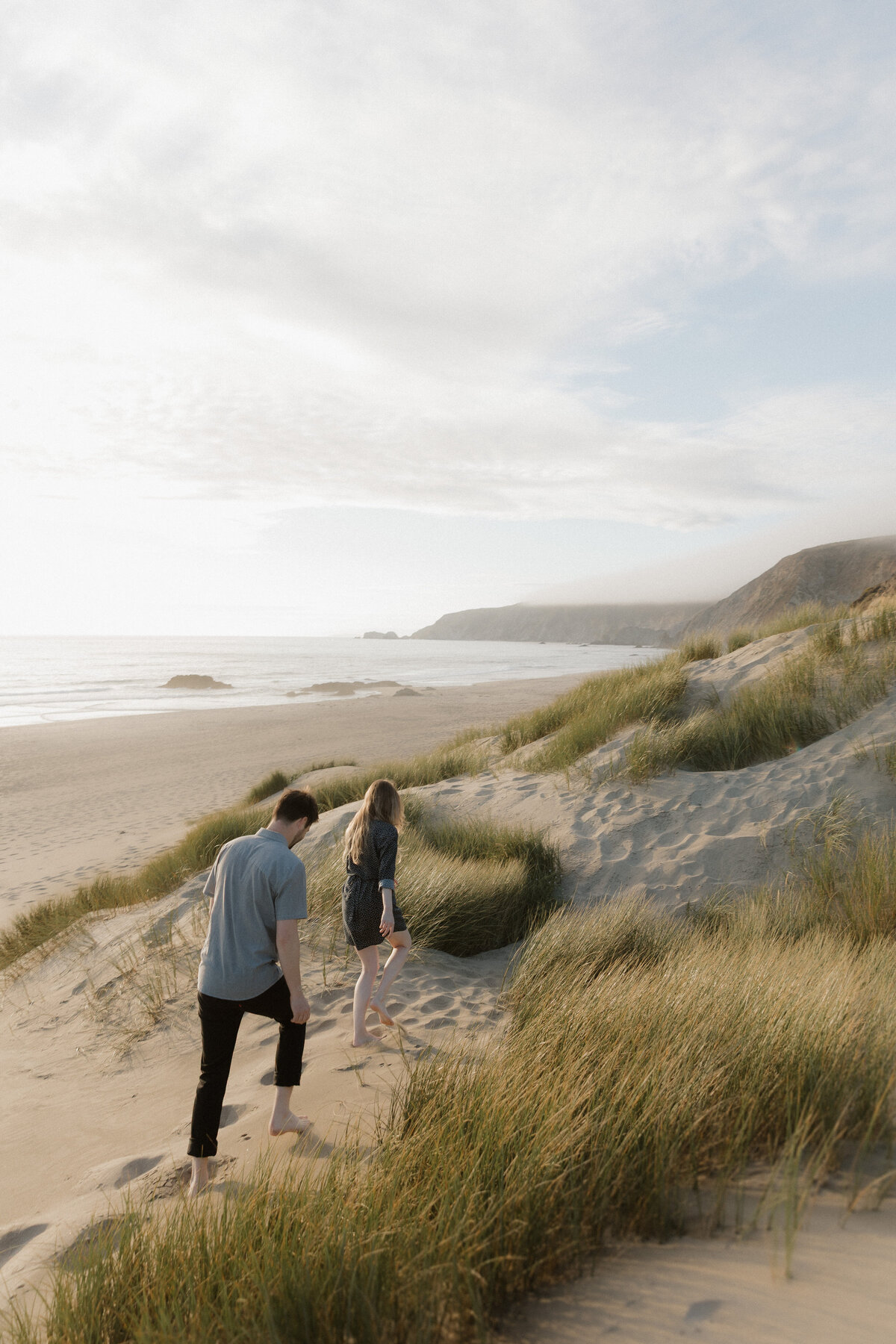 A couple walks barefoot through coastal sand dunes, embracing the beauty of natural, documentary-style wedding photography.