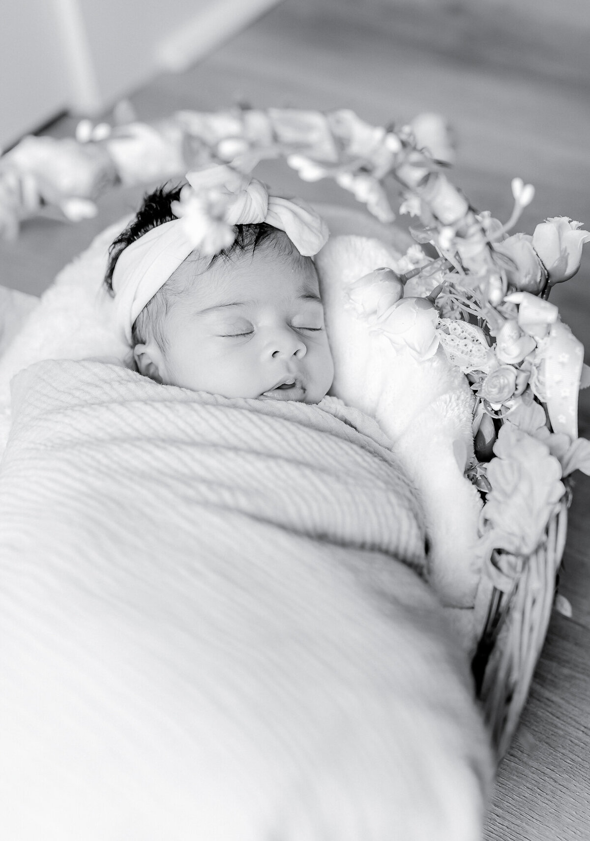 A newborn baby girl sleeps peacefully in a floral-adorned basket, wrapped in a soft blush swaddle with a delicate bow headband.