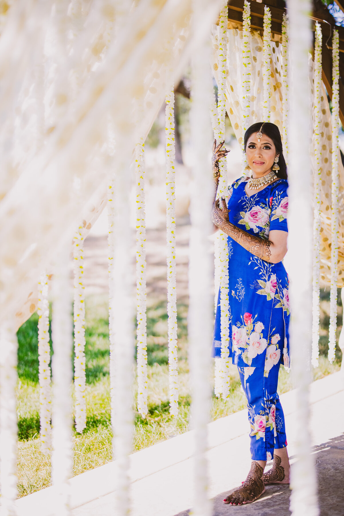 Sikh wedding bride during her mehndi day with fresh henna on her hands and feet posing near garlands in a floral blue outfit