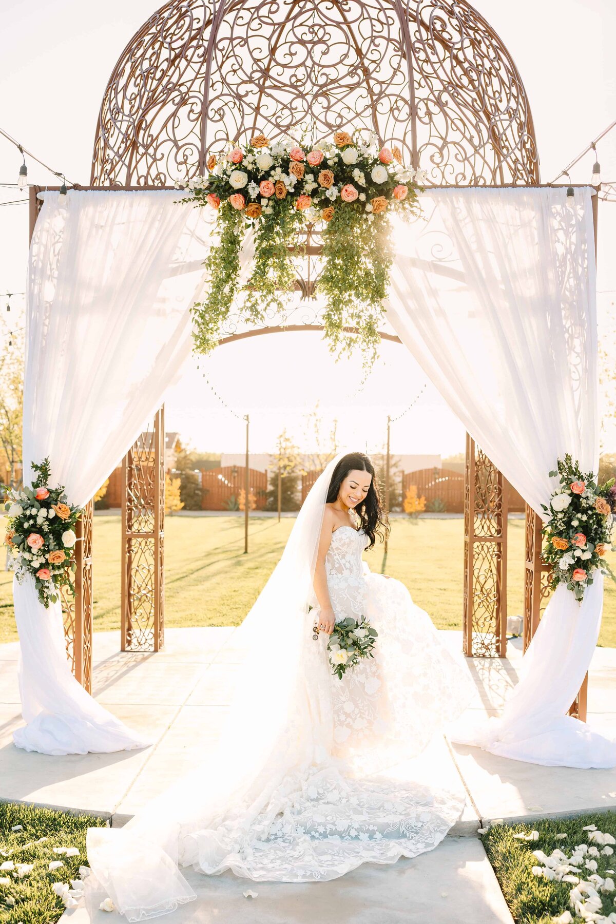 bride dancing in front of her wedding arch filled with stunning orange, coral, white florals, as the sun catches the  white drapery and her hair