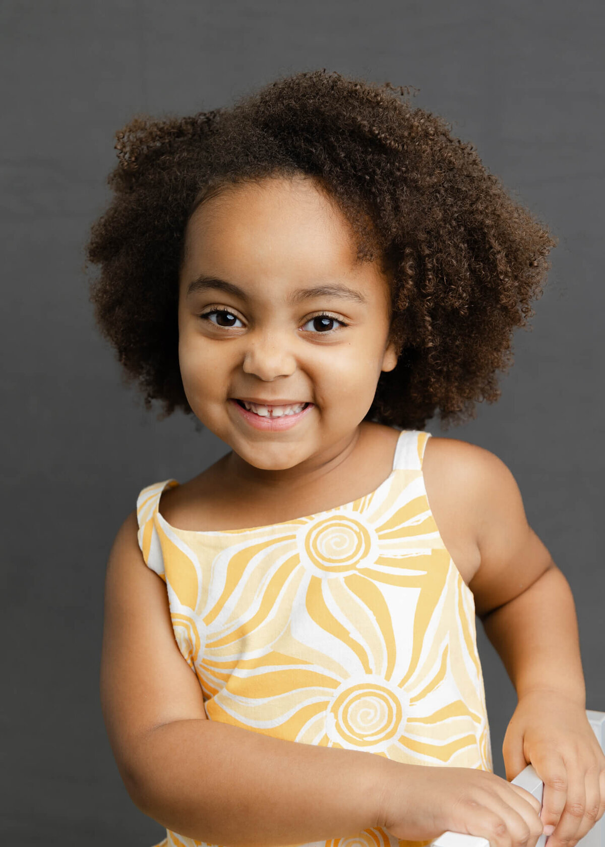 Black girl with curly hair in  a yellow and white sun shirt smiles sweetly for her portrait