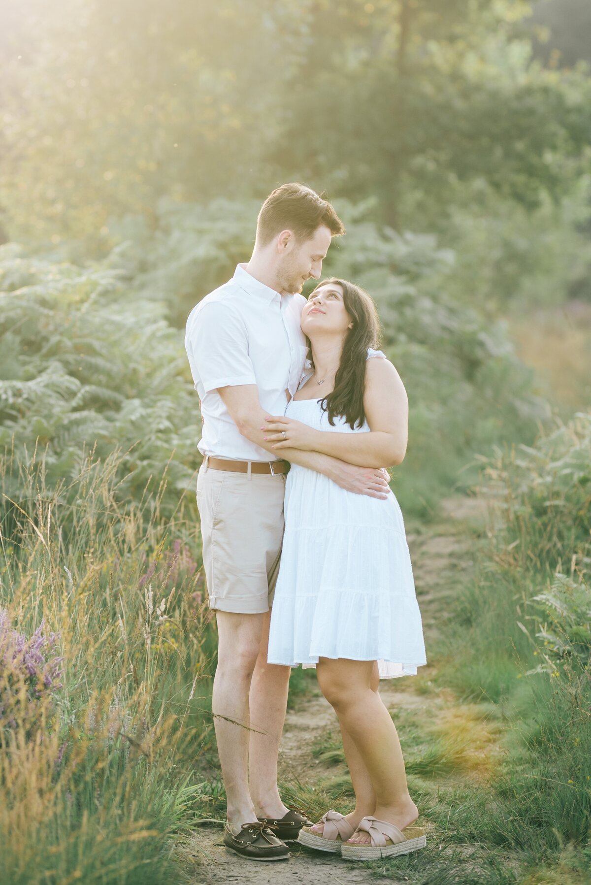 Engagement photoshoot, couple are cuddling at chobham cobham, she wears a white dress he wears a white shirt and beige shorts