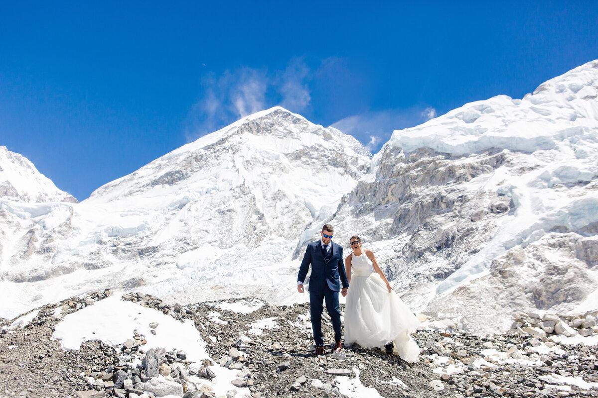The bride and groom, in gown and suit, go hiking through the rocky hills atop a glacier at Everest Base Camp for an adventure elopement in Nepal