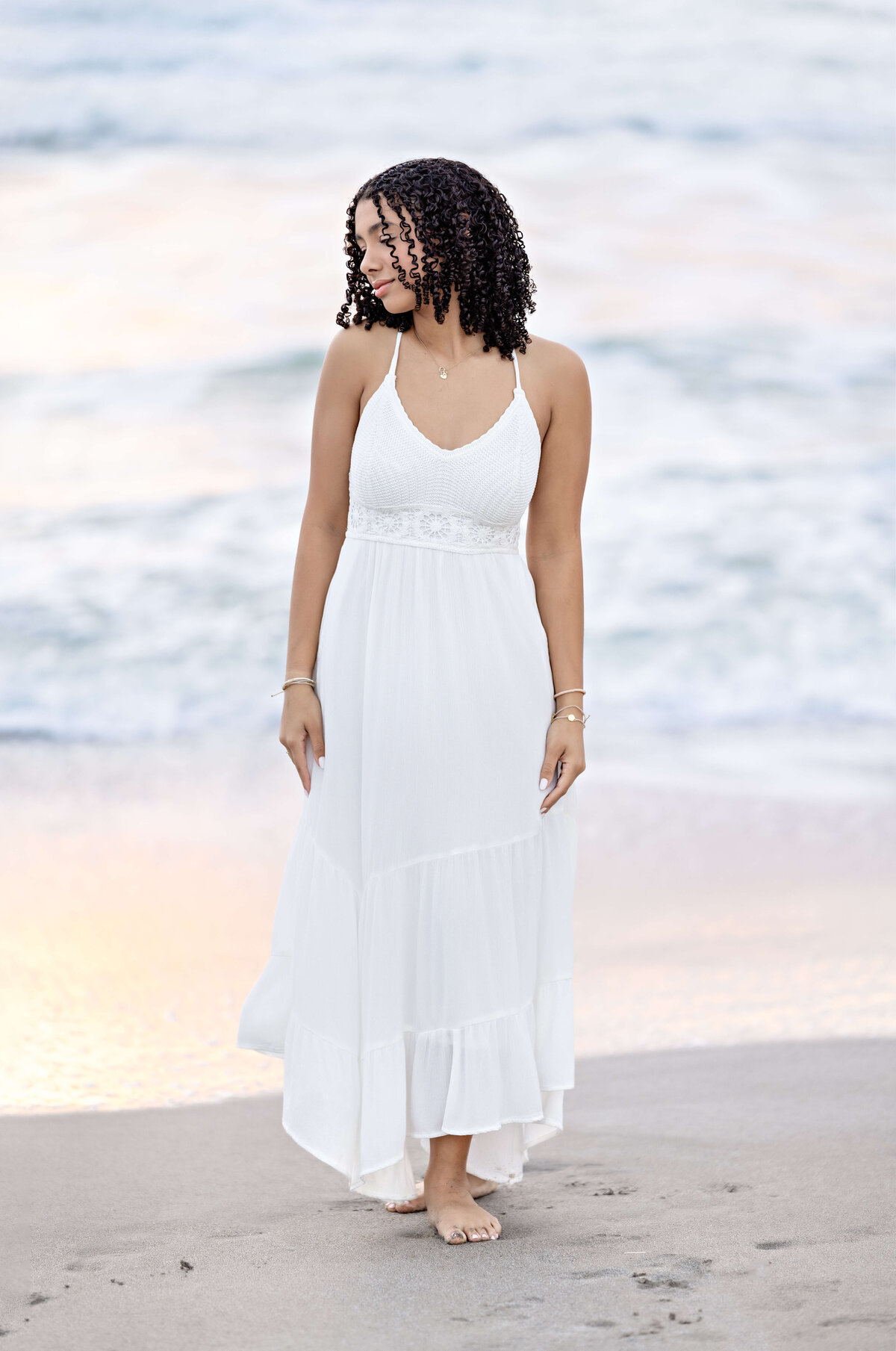 Senior girl looking down and off to the distance, softly fierce expression, white dress flowing with romantic energy at the beach.