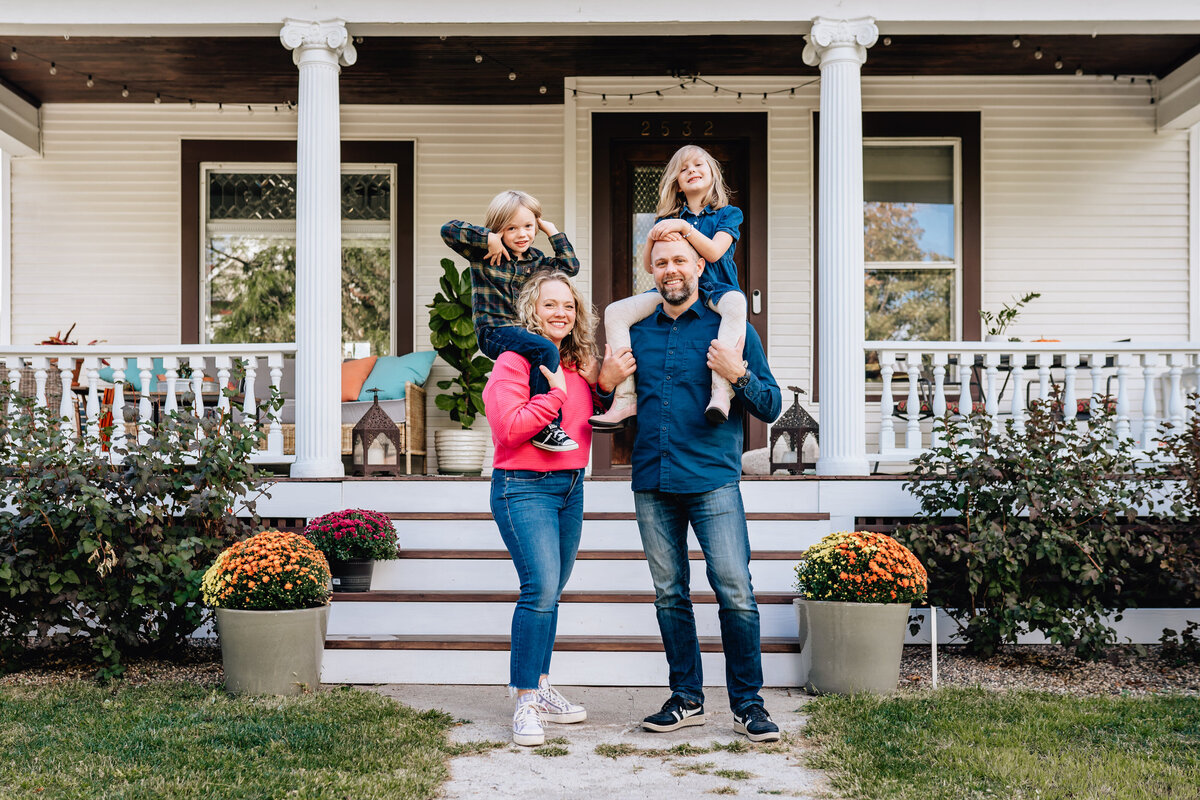 A family portrait in front of a house. The father and mother each carry one child on their shoulders, standing on a front path with white columns and greenery.